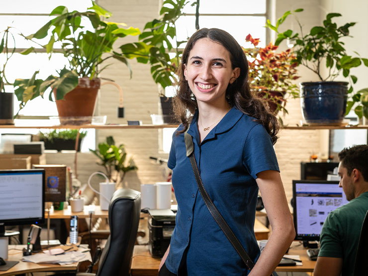 A young adult, in professional office attire, stands with hands in pockets in an office decorated with bright green plants.