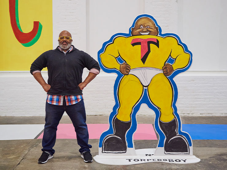 An artist stands, arms akimbo, in an gallery next two a life-size cutout of a super hero cartoon resembling the artist.