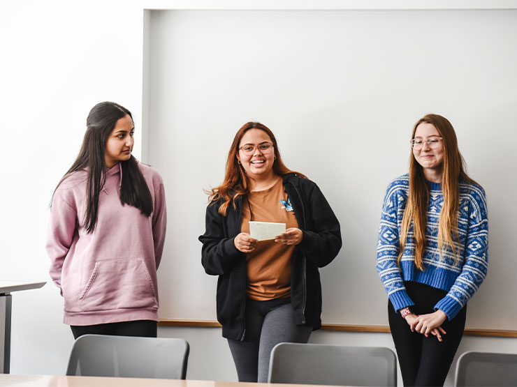 Three young adults stand against the wall with notes and deliver a presentation to a classroom.