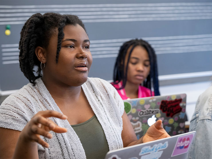 A young adult with braided hair speaks to someone off-camera in a classroom with an open laptop.