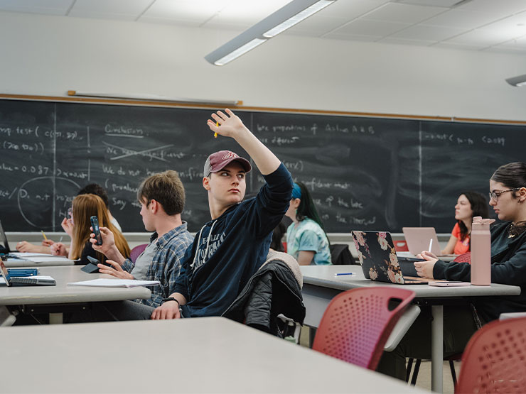 A college student wearing a baseball cap raises a hand in a classroom.