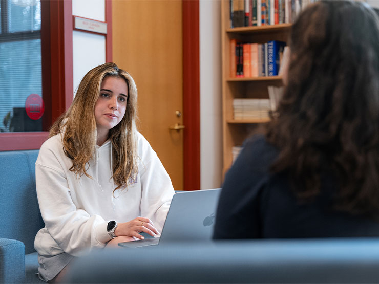 A college student sits at an open laptop and speaks with another student, just out of frame, on a pair of blue couches in the library writing center.