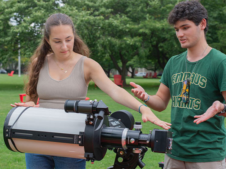 Two young adults stand outdoors and adjust a large telescope.