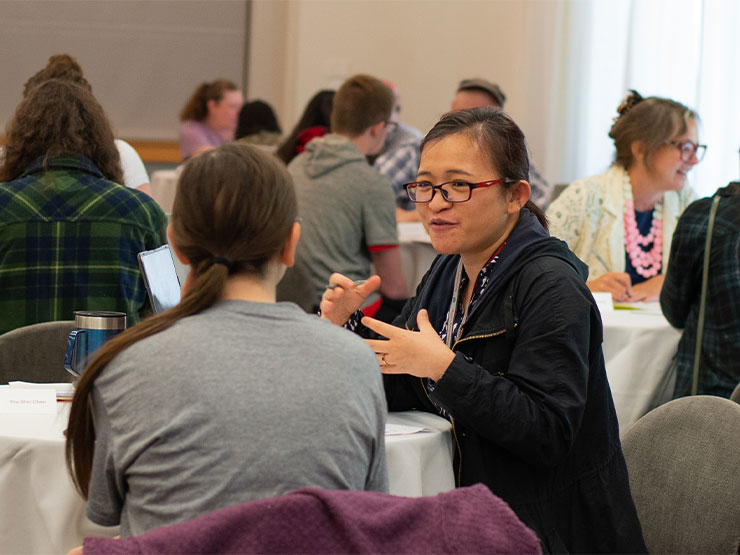 An adult and young adult seated at a table together speak while surrounded by similar pairings in a crowded room.