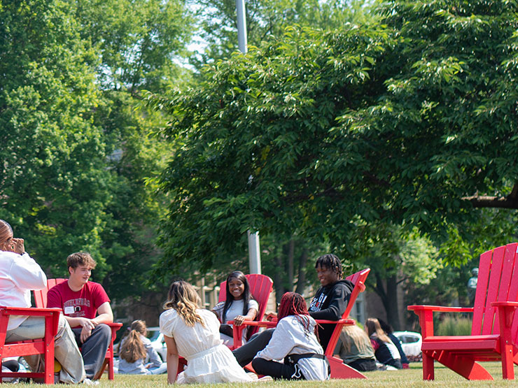 A group of students talk while sitting in red Adirondack chairs and on a green lawn on a college campus.