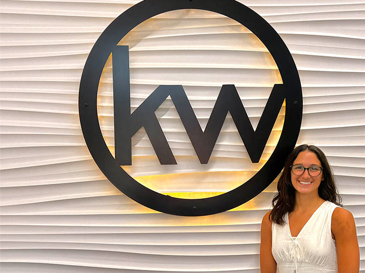 A smiling college students stands in front of a wall displaying the corporate logo for Keller Williams Real Estate.