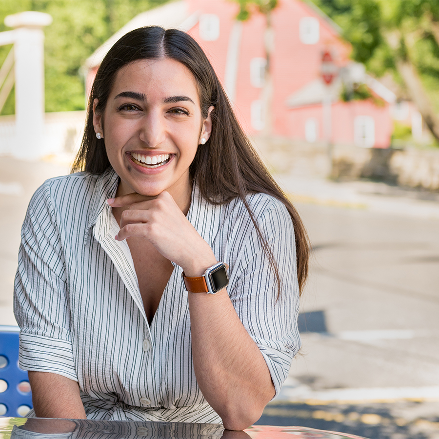 A woman, seated outdoors near a water mill, smiles with her hand beneath her chin.