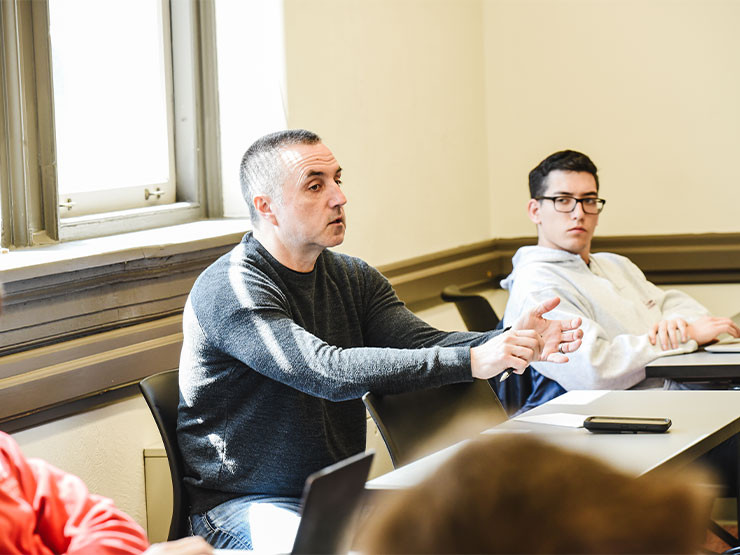 An instructor speaks while sitting in the back of the classroom, flanked by two students at desks.