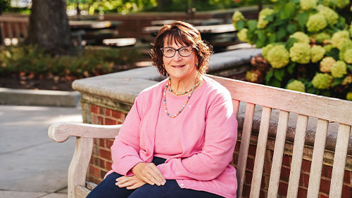 A woman in a pink sweater and glasses poses for a photo on a bench