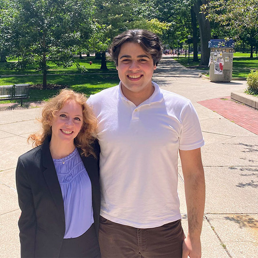 A pair of young adults outdoors smile at the camera, one arm around each other.