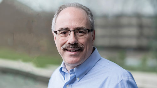 A man wearing glasses and a light blue dress shirt smiles at the camera.