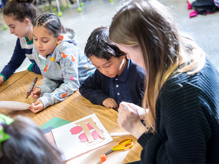 A young adult helps a group of elementary school students seated around a table create crafts.