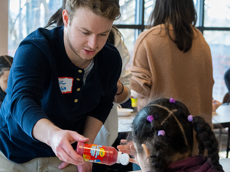 A young adult pours red paint into a bowl for elementary school children seated at a desk in a classroom.