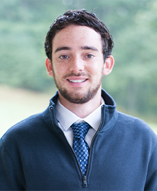 An adult, wearing a blue pull over with a dress shirt and blue tie beneath, smiles at the camera.