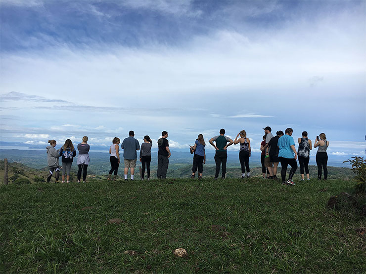 A group of 15 young adults stand, with backs to the camera, on summit, with land and sky spread out before them.