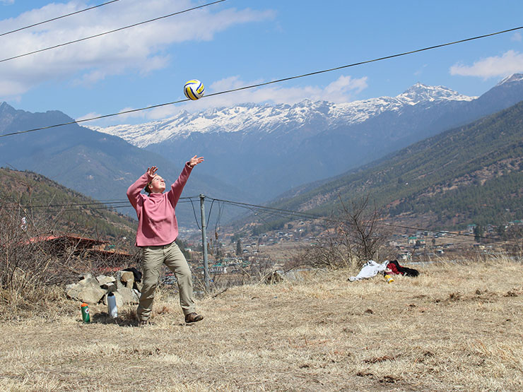 An college student in a pink sweater prepares to strike a volleyball in a grassy field with a dramatic view of snow-capped mountains in the distance.