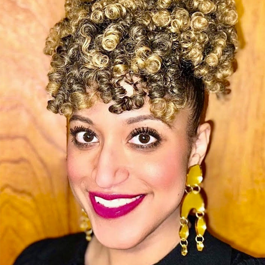 A woman with curly hair, pink lipstick and gold earrings smiles at the camera.