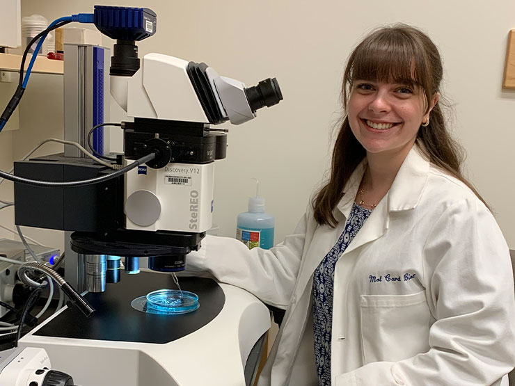 An adult with long brown hair and bangs wear a white lab coat while siting at a microscope and smiling at the camera.