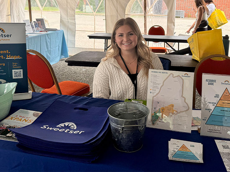 College student Greta Marchildon smiles while seated behind a table at mental health awareness event.