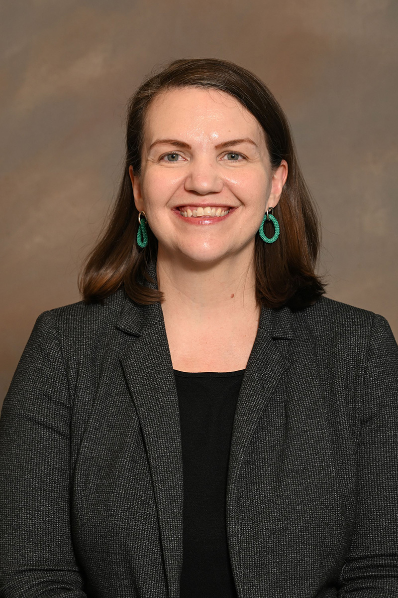 Portrait image of professor, Sarah Runcie, seated wearing dark grey cardigan and black shirt.