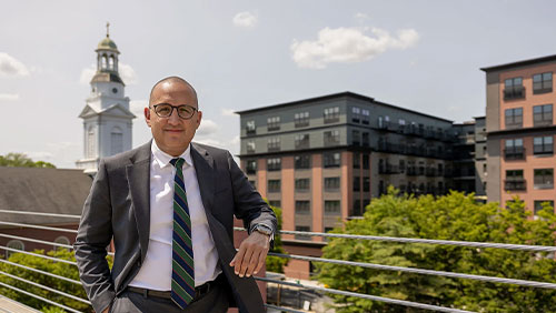 A man in a suit and glasses poses against a railing overlooking a city.