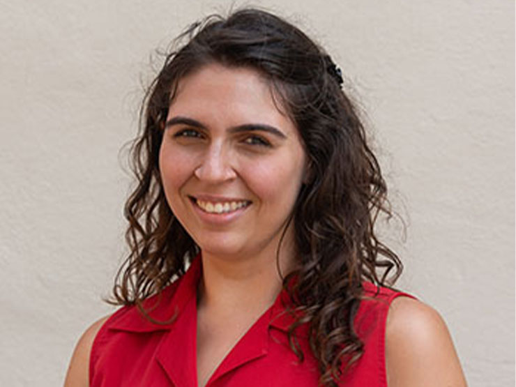 A woman with curly shoulder-length hair in a red sleeveless blouse smiles at the camera.