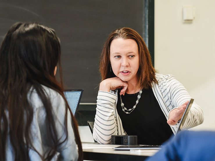 A college professor speaks with a students and points to the screen of a laptop in a classroom.