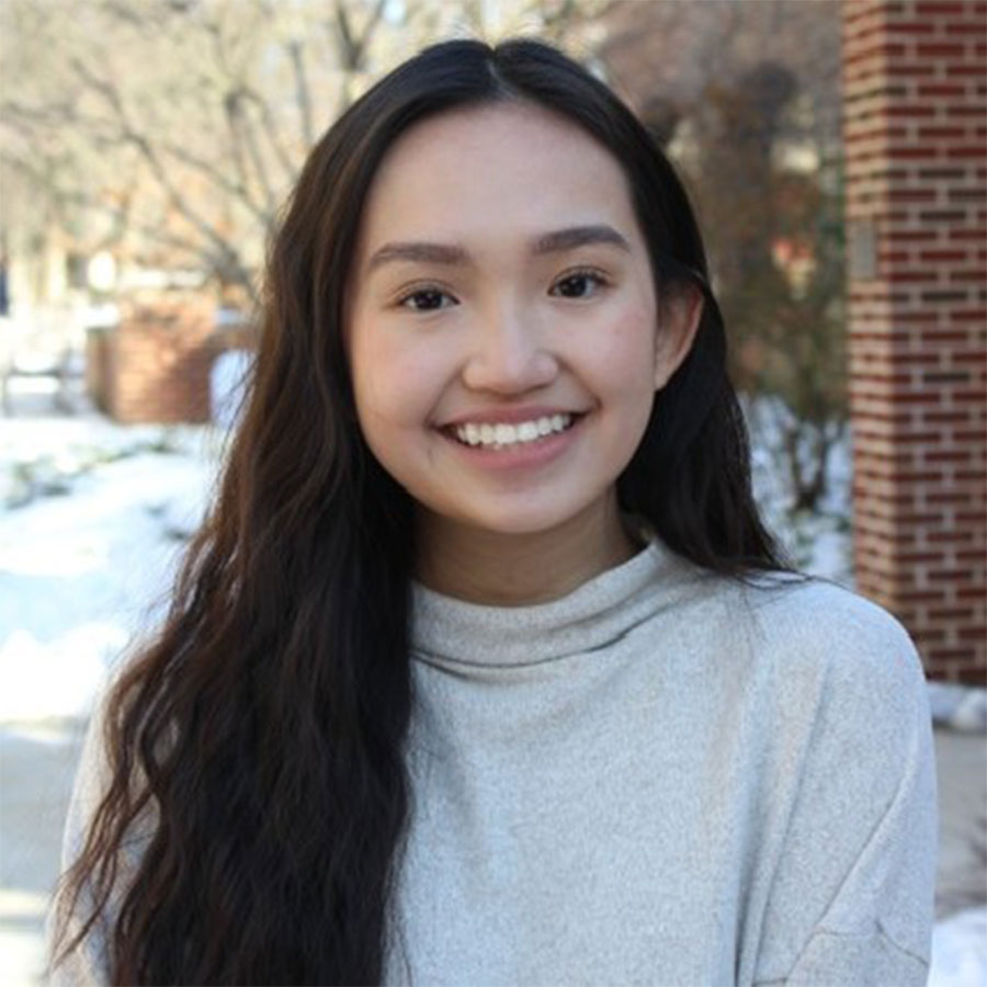 A young adult with long black hair wears a white turtleneck outdoors and smiles at the camera.