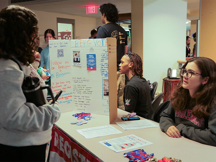 A college student involved with the nonpartisan group BergVotes sits at an informational table and speaks to another student.