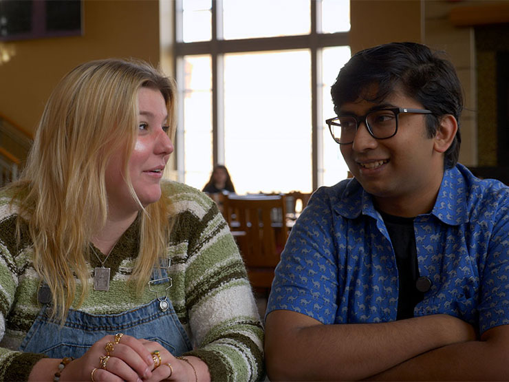 A pair of college students being interviewed turn to speak to one another while seated in a dining hall.