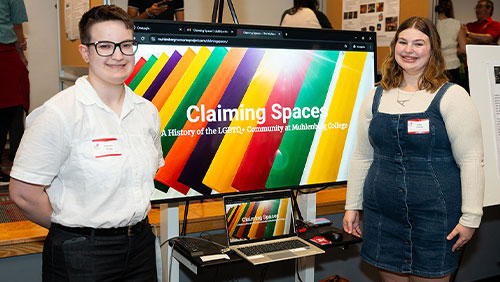 Two college students stand in front of a video screen presentation; text on the screen says Claiming Spaces