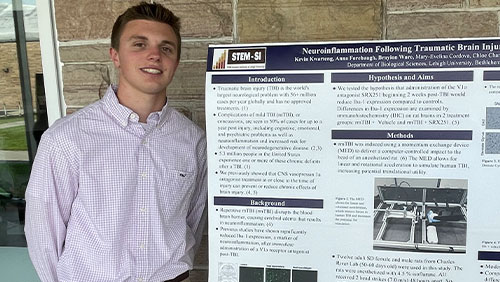 A college student wearing a dress shirt and slacks smiles while standing next to a large poster board with details about a research project.