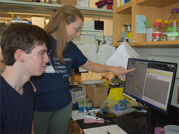 A college instructor works with a student in a lab, pointing to data on a computer screen.