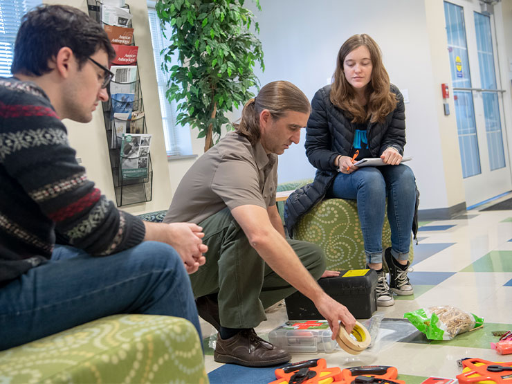 An instructor with a long brown ponytail sorts items on the floor of learning space while students look on.