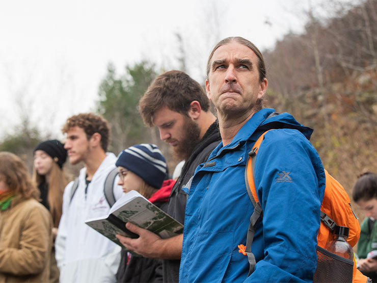 An instructor, wearing a blue jacket and orange backpack looks into the distance while outdoors with a group of students.