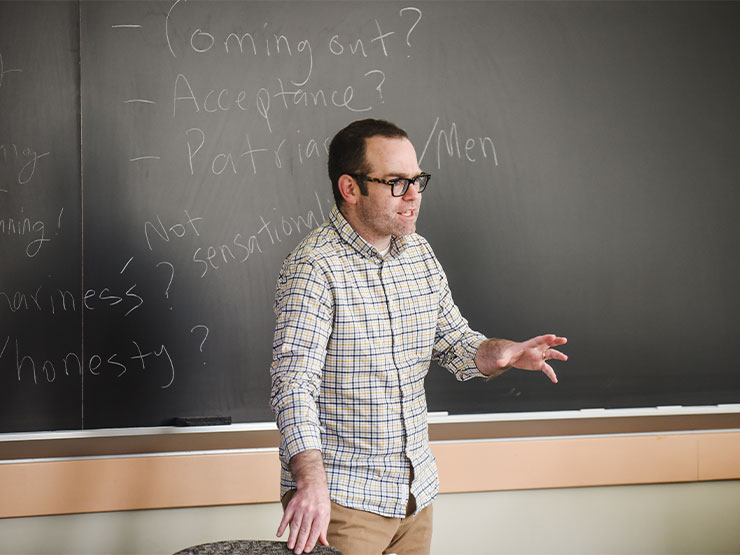 An instructor speaks to a class while standing in front of a chalkboard.