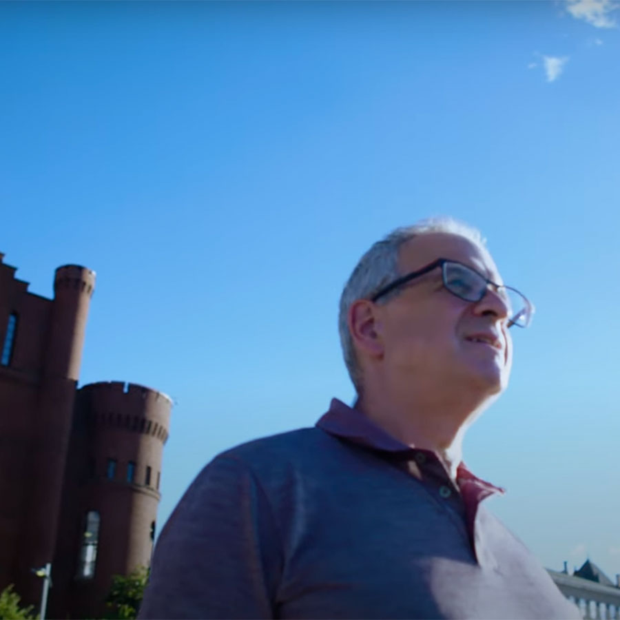 A man in a blue shirt and glasses looks off into the distance against the backdrop of a bright, blue sky.