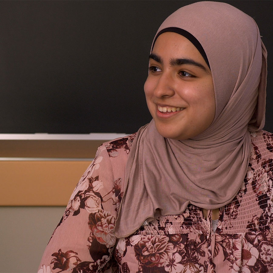 A college students wearing a beige headscarf and floral blouse smiles at someone off-camera.