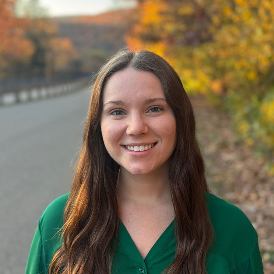 A young person in a green shirt smiles for a photo outside with fall foliage in the background