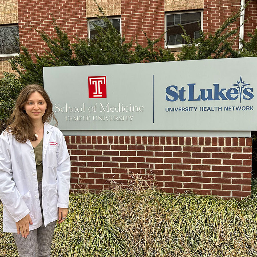 A medical student wears a medical coat and stands outside of the Temple School of Medicine and St Lukes Hospital.