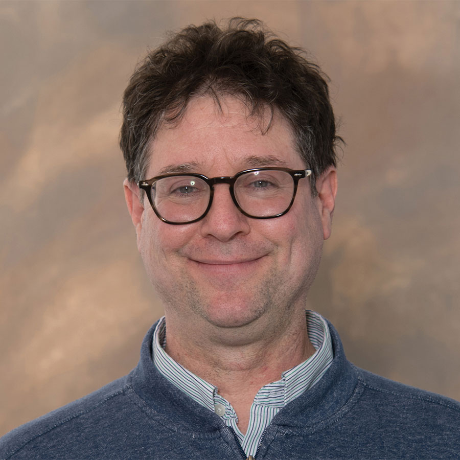 A professional headshot of a smiling man with glasses and a blue sweater pullover.