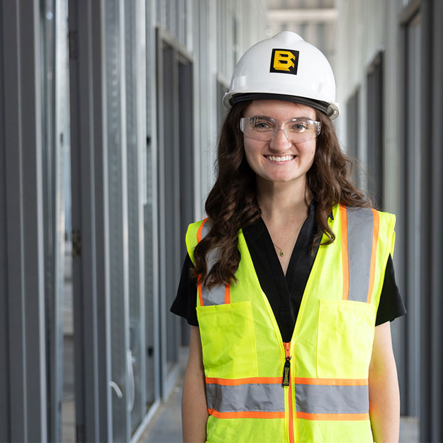 Brianna Moyer smiles with a hard hat and neon vest, surrounded by metal girders in a construction site.