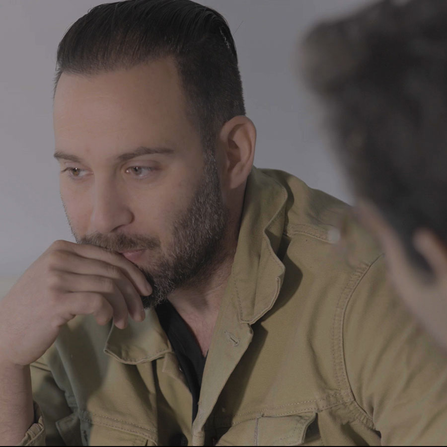 A still image from a film where a man in an olive-colored shirt sits with his hand resting at his chin in a thoughtful expression.