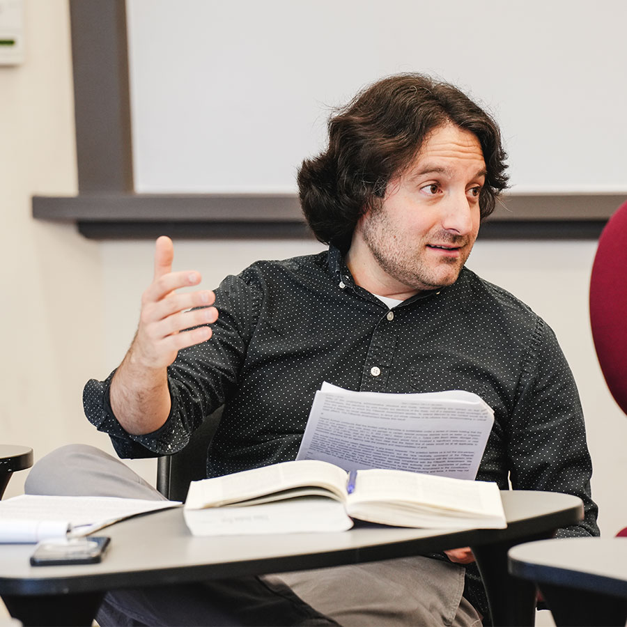 A college professor sits at a desk and speaks to a classroom.