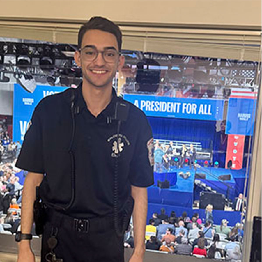 A college student in an EMS uniform stands in front of a window overlooking a rally stage