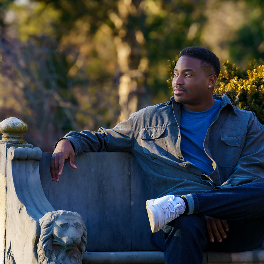 A man in a jacket sits outdoors on a stone bench in the fading light of day.