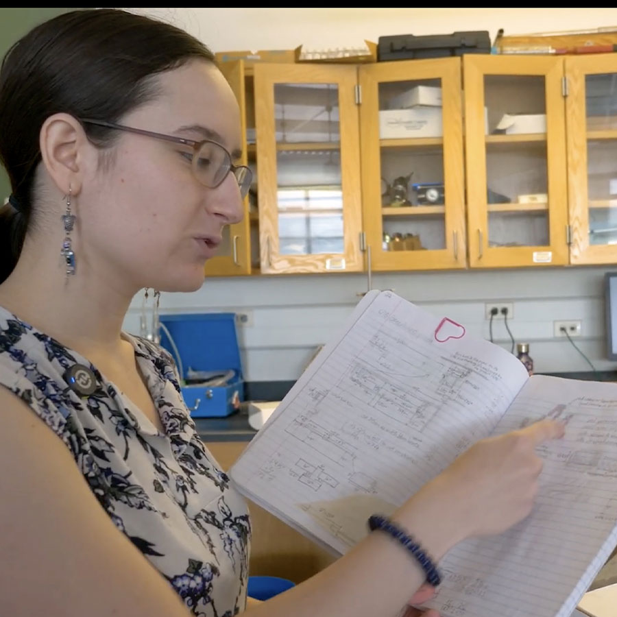 A college student speaks while referencing an open notebook in a lab.