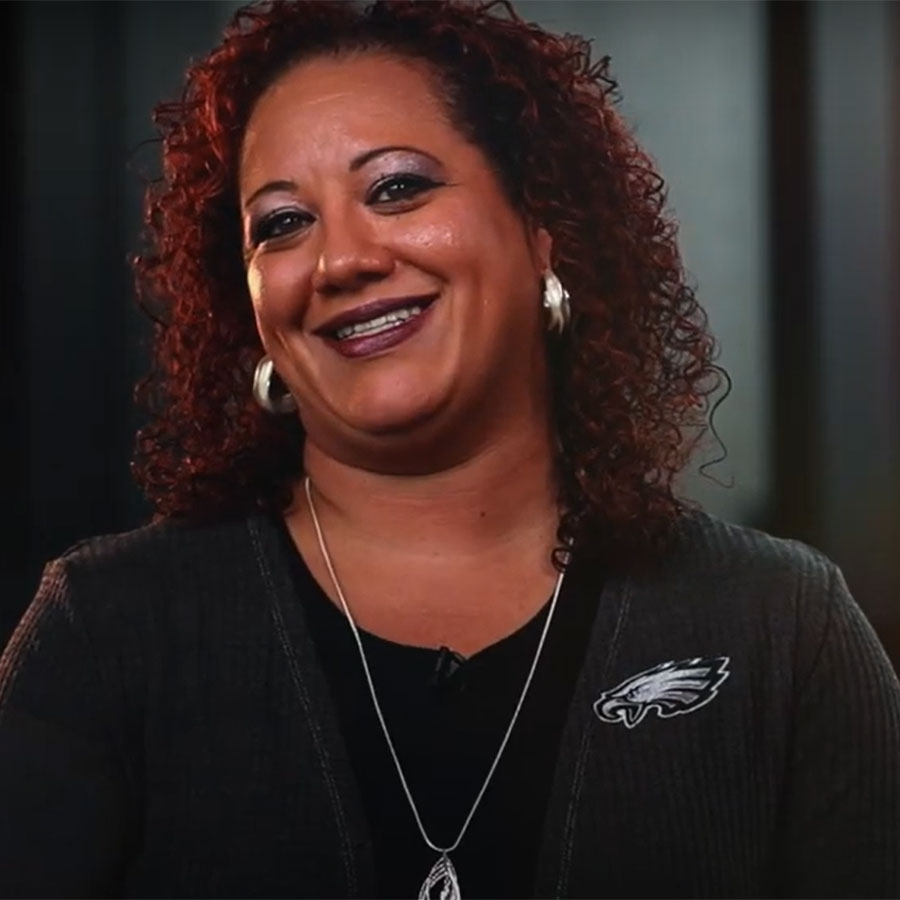 An adult with curly shoulder-length hair, wearing a sweater with a Philadelphia Eagles logo smiles at the camera.