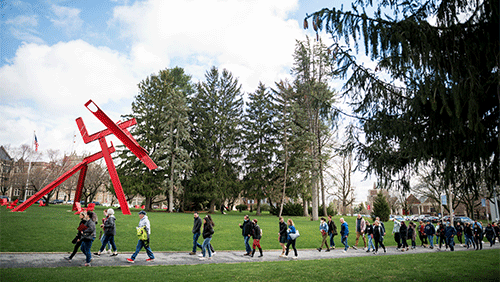 Clusters of families with young adults walk along a walkway on Muhlenberg College campus near the red modern sculpture Victors Lament.