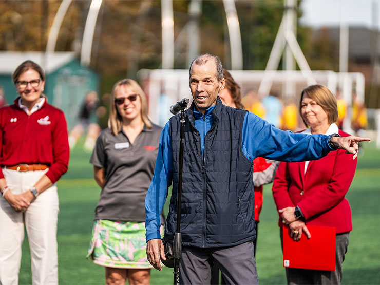Alumni Jeff Morris speaks into a microphone on an athletic field with Muhlenberg officials standing behind him.